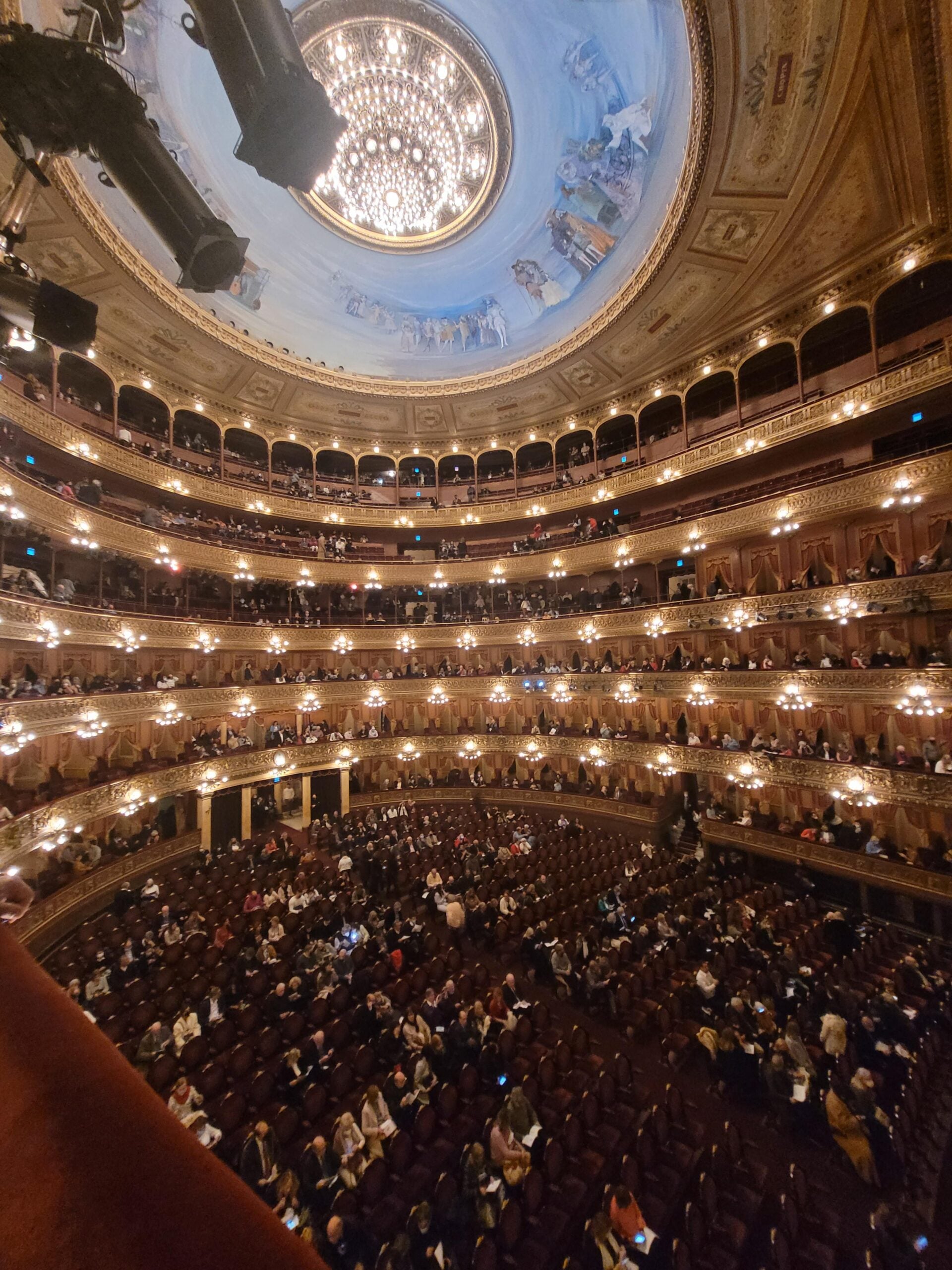 A grand opera house interior with ornate balconies, warm lighting, and a large, decorated dome ceiling. The audience is seated, with some empty seats still visible. The view is from one side of the theater.