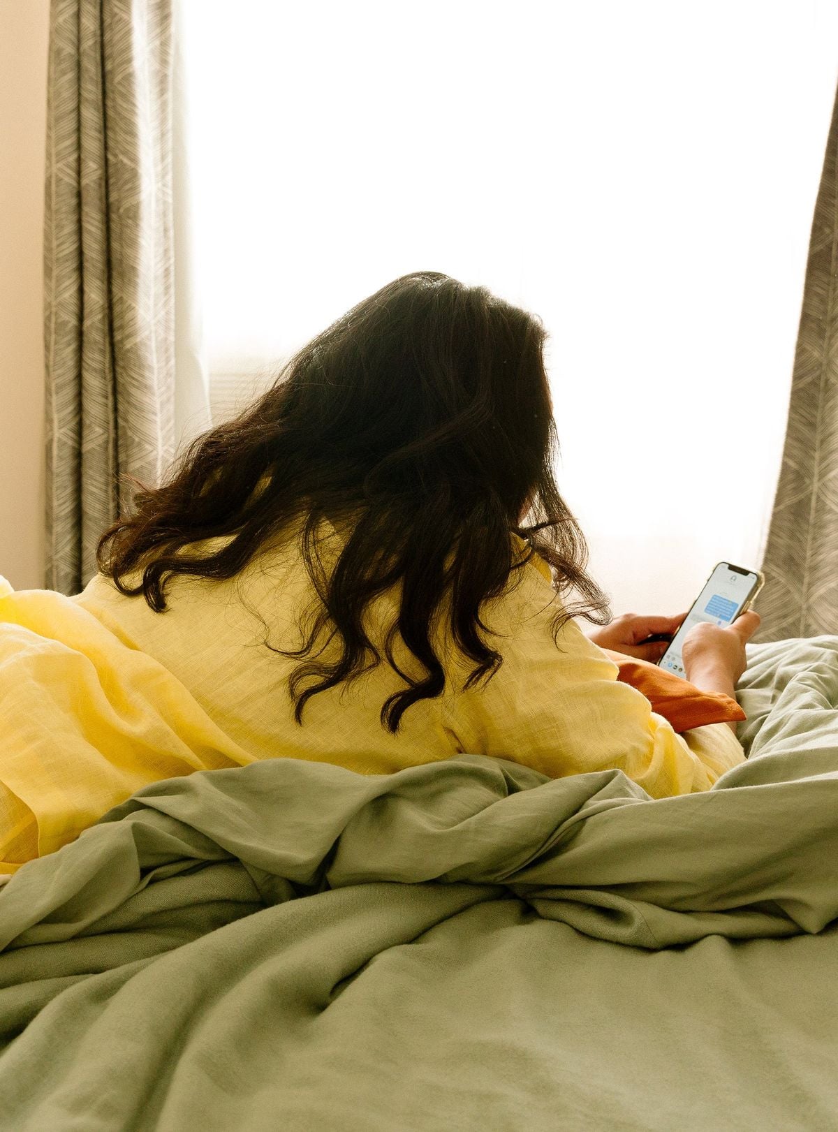 A person with long dark hair, wearing a yellow top, lies on a bed with green sheets and looks at a smartphone. Light enters the room through a window with patterned curtains in the background.