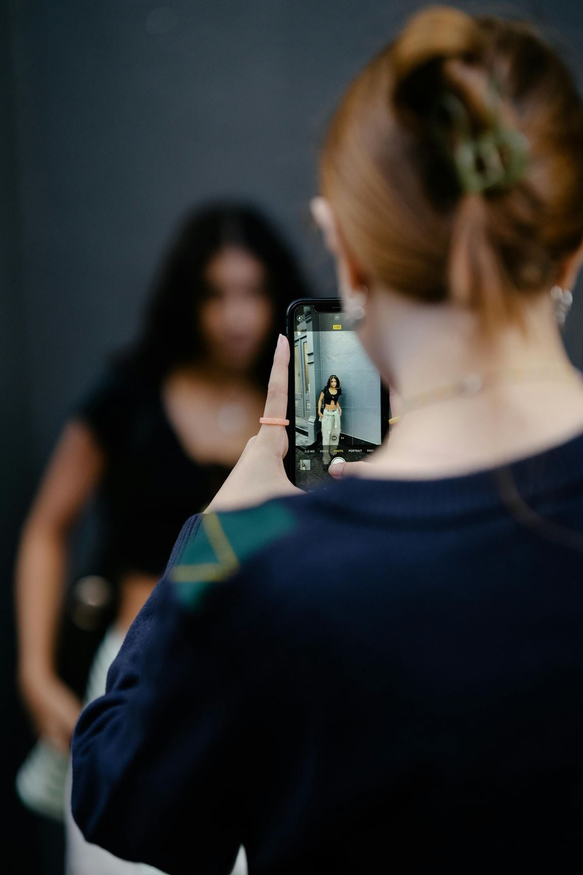 A person with reddish hair in a clip holds up a phone to take a photo of another person standing in the background. The phone screen shows the subject posing for the picture.