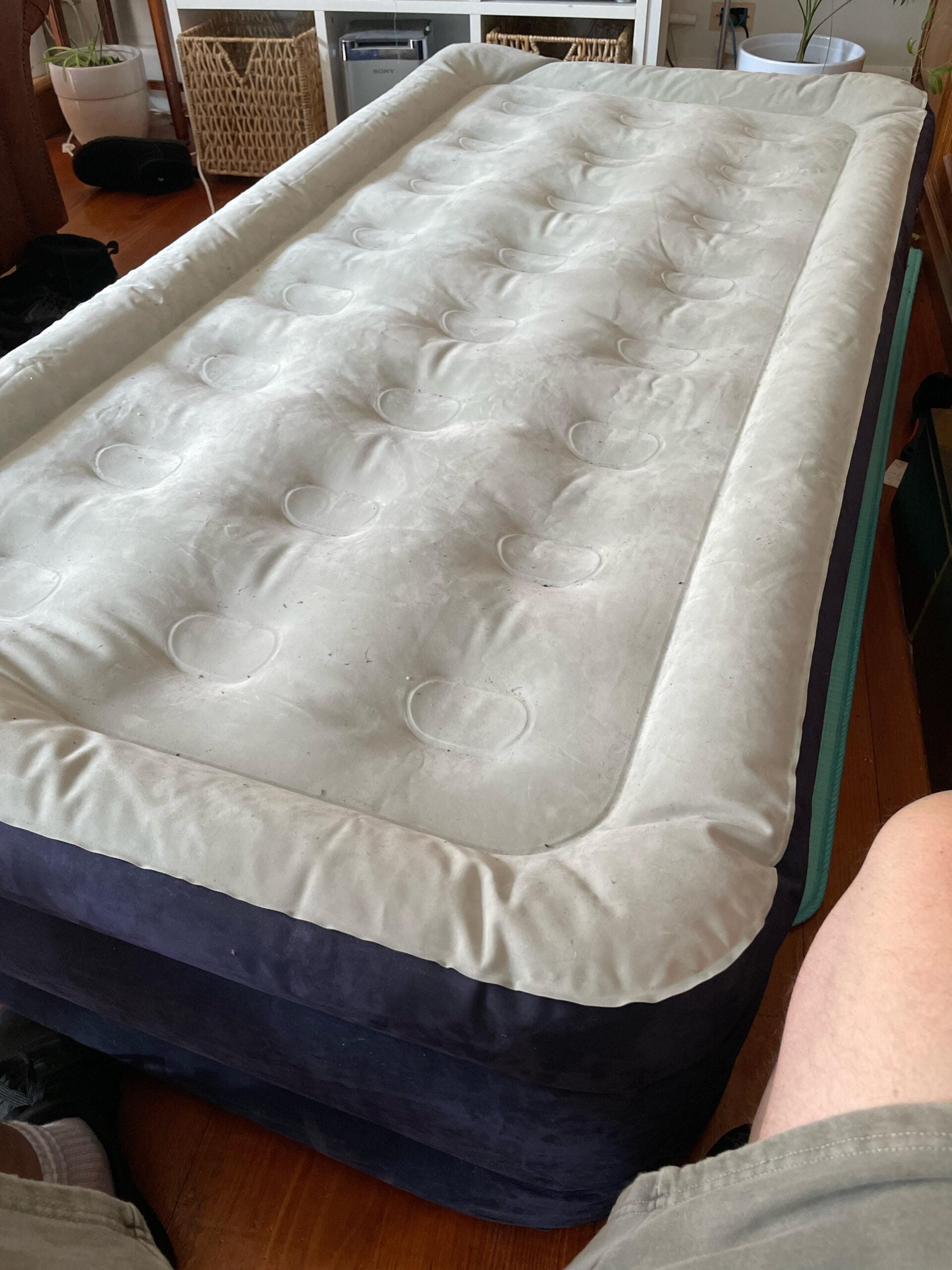 An inflated air mattress with a light beige top and dark blue sides sits on a wooden floor. Storage baskets and furniture are visible in the background, and a person's legs are partially visible in the foreground.
