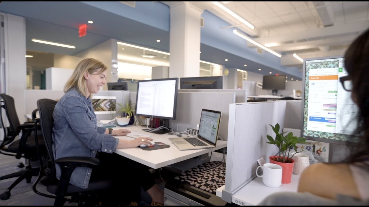 A woman is smiling while working at a computer desk in a modern open office. Another person is partially visible at a nearby desk with a monitor, plants, and a coffee mug. The office is bright and spacious.