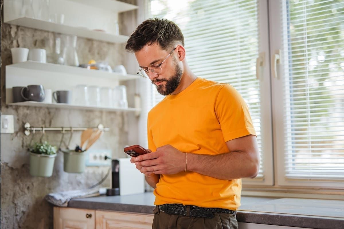 A man in a bright orange t-shirt stands in a kitchen by a window with blinds, looking at and using his smartphone. Shelves with dishes and potted plants are visible in the background.