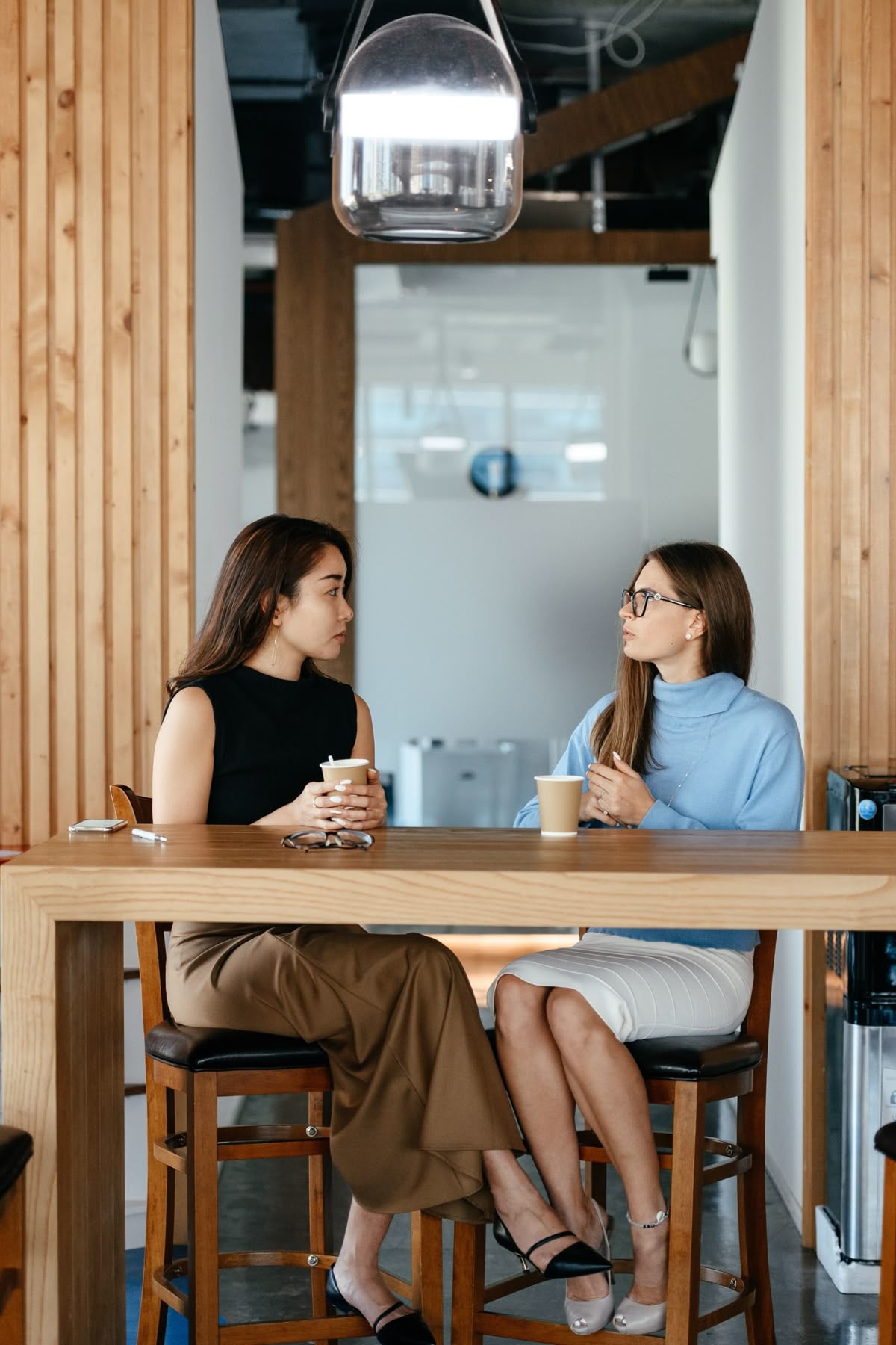 Two women sitting on high chairs at a wooden table indoors, holding coffee cups and having a conversation in a modern office or café setting.
