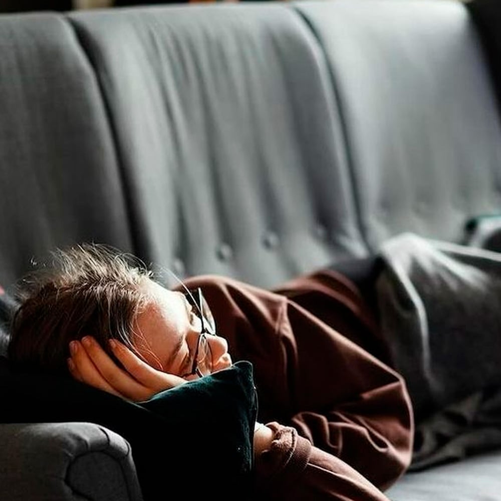 A person wearing glasses rests on a gray couch, lying on their side with their head on a dark pillow, partially covered by a blanket.