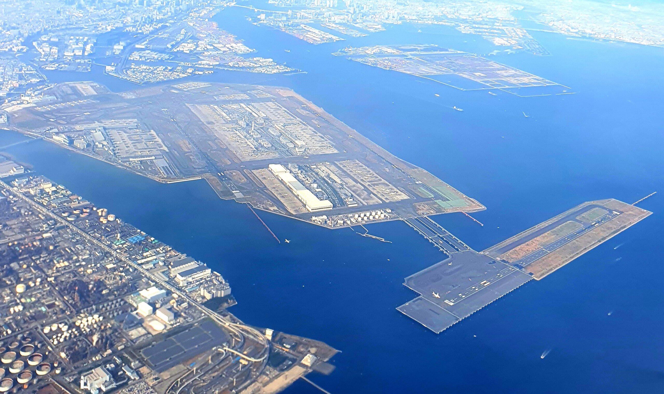Aerial view of an airport with runways extending over water, surrounded by city buildings and coastline. Multiple runways, taxiways, and terminals are visible, with blue ocean and distant urban areas in the background.