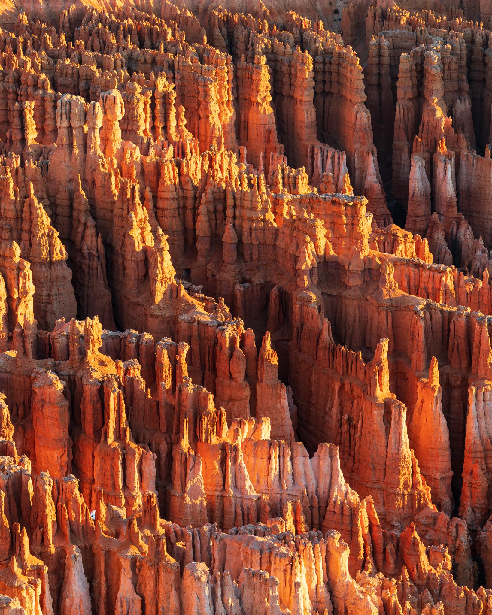 Tall, narrow, orange-red rock formations rise closely together under bright sunlight, creating dramatic shadow patterns in a rugged, canyon-like landscape.