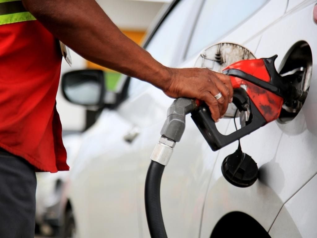 A person in a red shirt is refueling a white car at a gas station, holding a black and red fuel pump nozzle inserted into the car’s fuel tank.