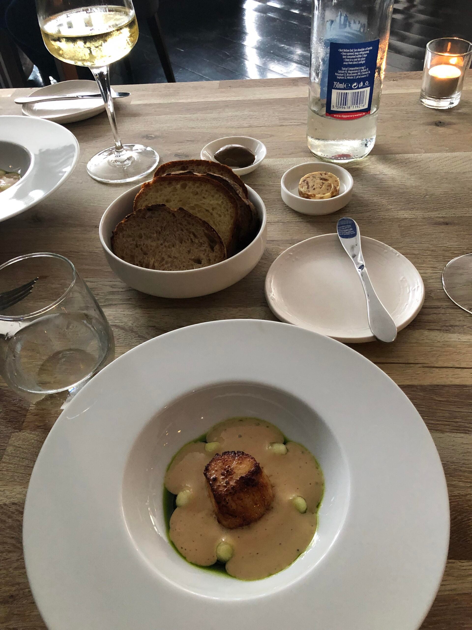 A plated dish with a seared scallop in a creamy sauce, garnished with green oil and dots of puree. There’s a bowl of sliced bread, butter, a glass of white wine, bottled water, and a lit candle on a wooden table.