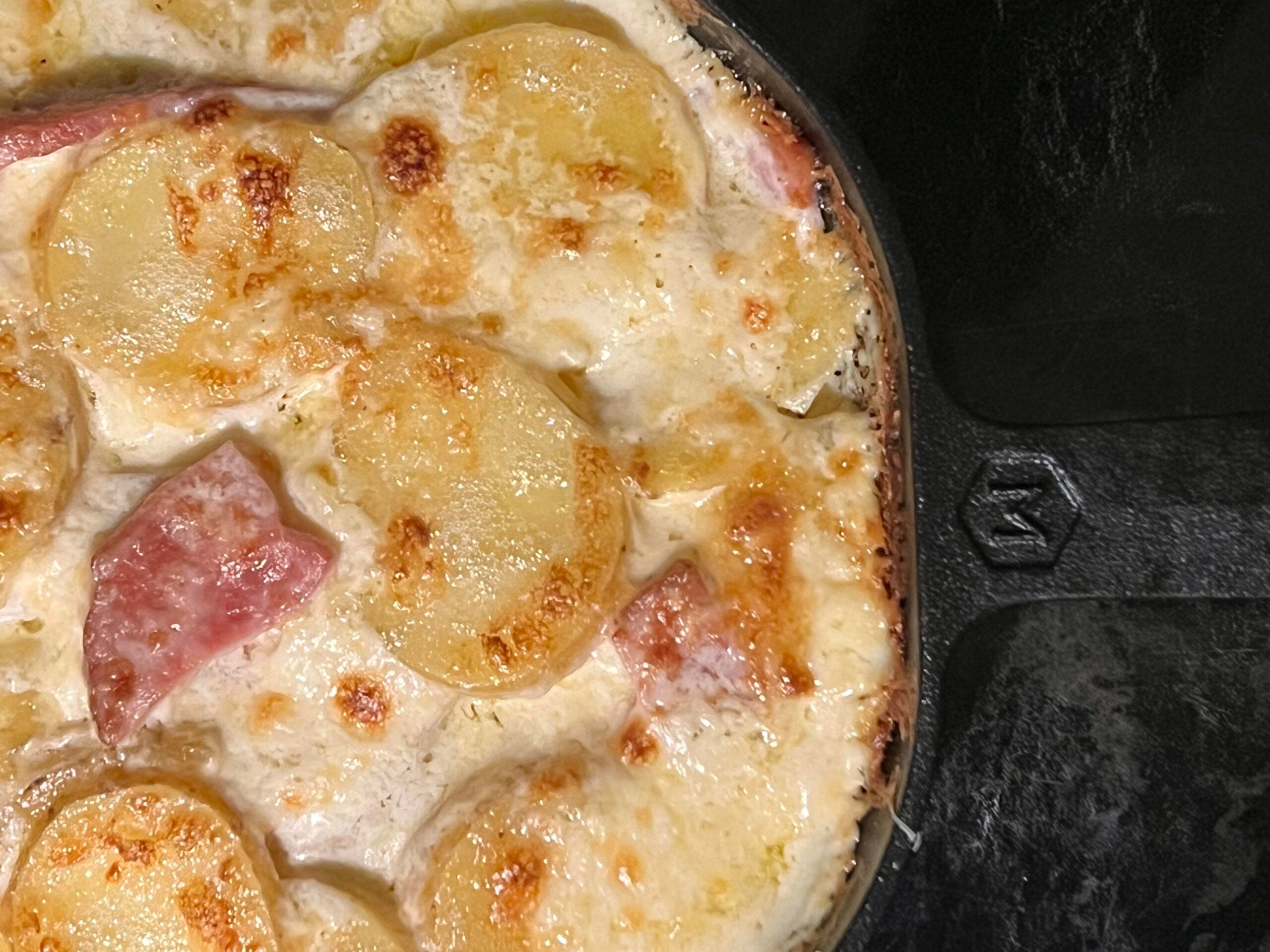 Close-up of a baked potato gratin in a cast iron skillet, featuring golden, crispy slices of potato with creamy sauce and pieces of ham, with some browned spots on top.