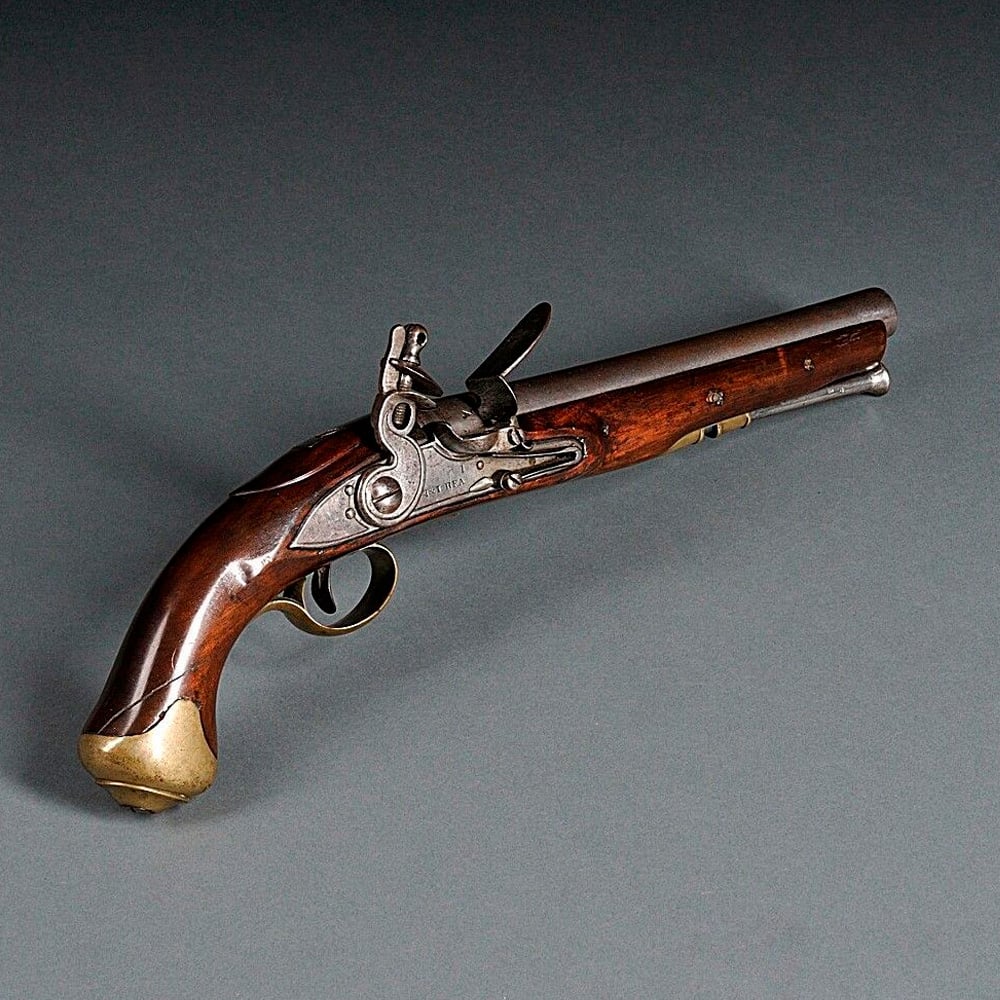 A vintage flintlock pistol with a wooden handle, brass butt, metal trigger guard, and steel barrel, displayed on a plain dark background.