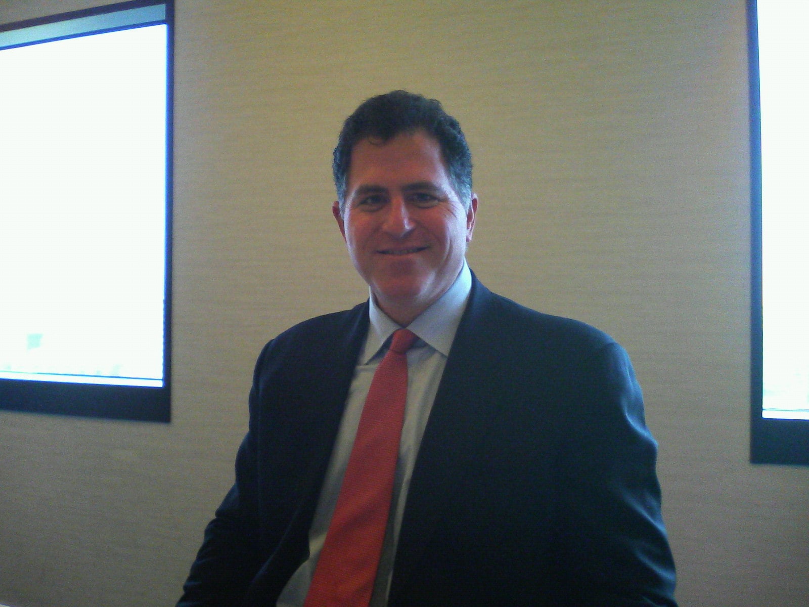 A man in a dark suit and red tie sits indoors, smiling at the camera. Two rectangular windows with bright light are visible behind him on a beige wall.