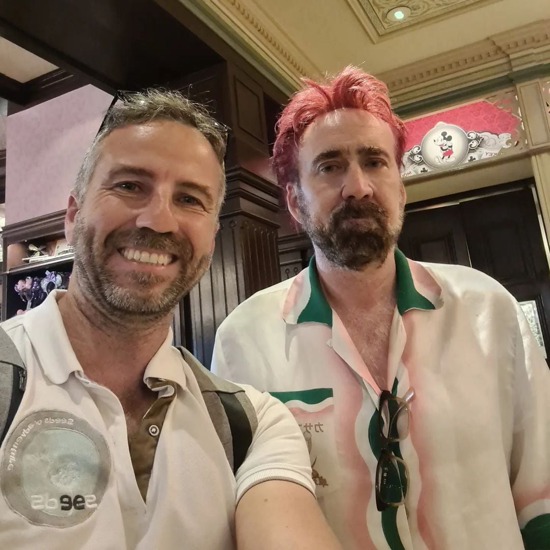 Two men stand indoors and pose for a photo together. The man on the left smiles at the camera, while the man on the right, with pink hair and a trimmed beard, has a neutral expression and wears a light-colored shirt with green accents.