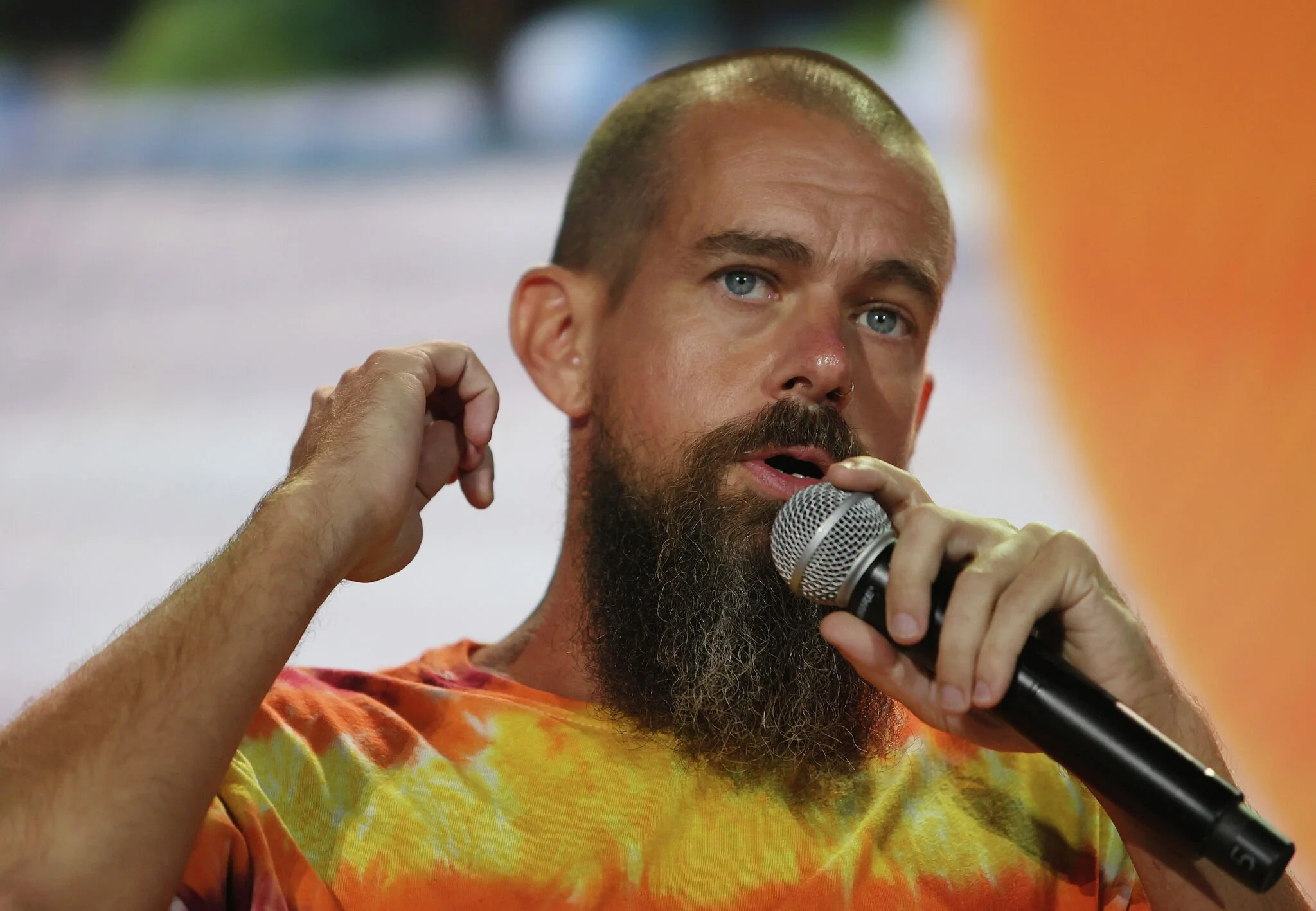 A man with a long beard and shaved head, wearing a tie-dye shirt, speaks into a microphone on stage, gesturing with his other hand. The background is blurred with orange and green hues.