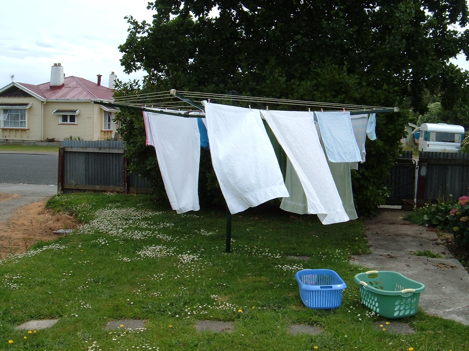 A rotary clothesline with towels and clothes hanging to dry in a grassy yard. Two empty laundry baskets sit on the ground nearby. A house, fence, and trees are visible in the background.