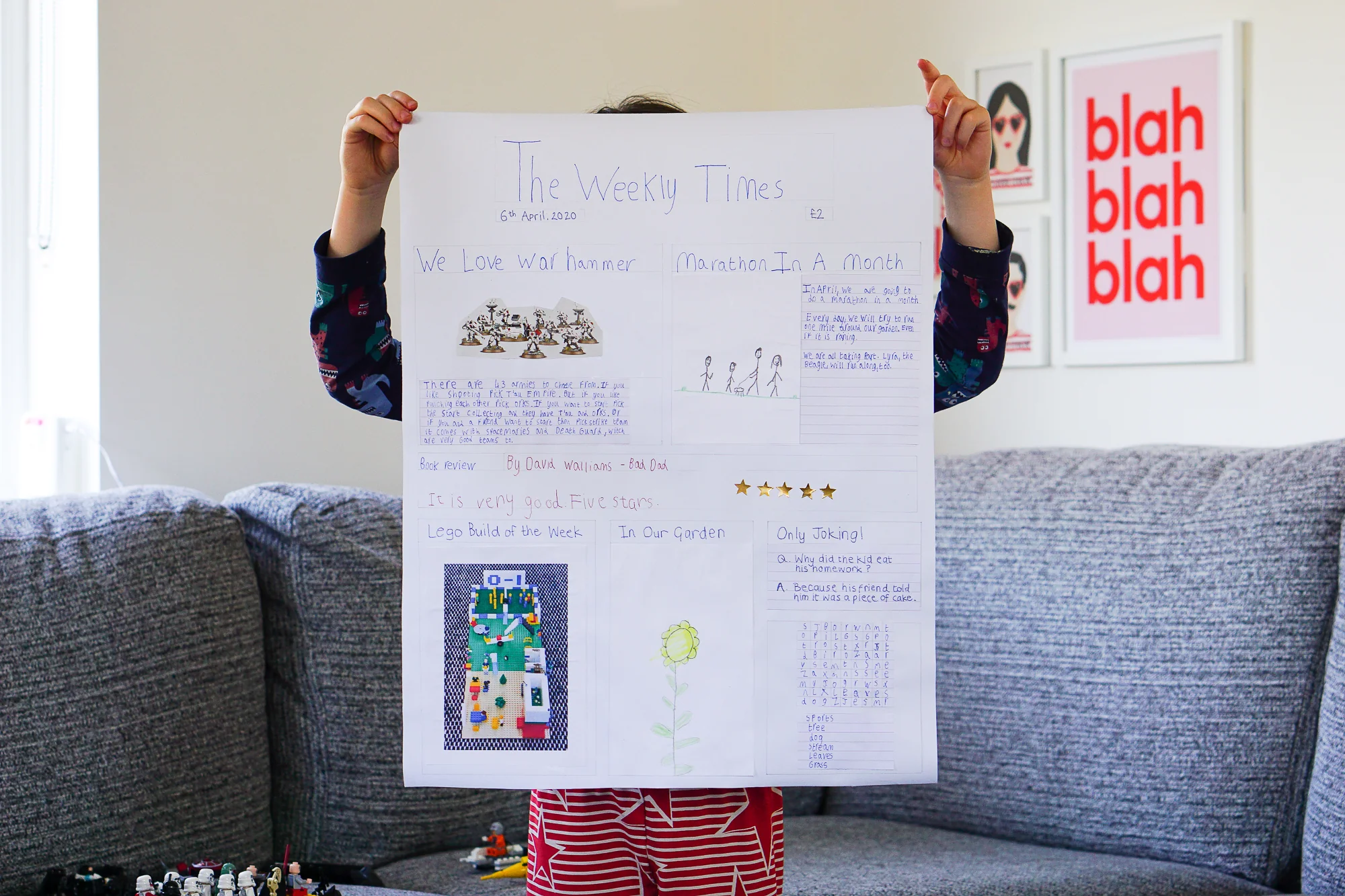 A child in pajamas stands on a couch holding up a large handmade newspaper poster titled "The Weekly Times," featuring drawings, handwritten text, and pictures. The child’s face is blocked by the poster.