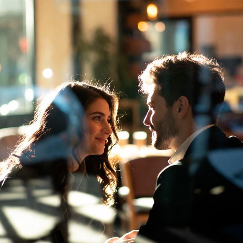 A woman and a man sit closely together at a table in a sunlit cafe, smiling at each other through a window that reflects light and shapes from outside. The atmosphere appears warm and intimate.
