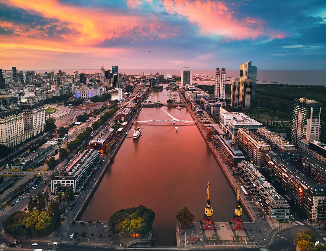 Aerial view of a cityscape at sunset with colorful clouds, showing a canal lined with modern and historic buildings, bridges, and greenery along the water.