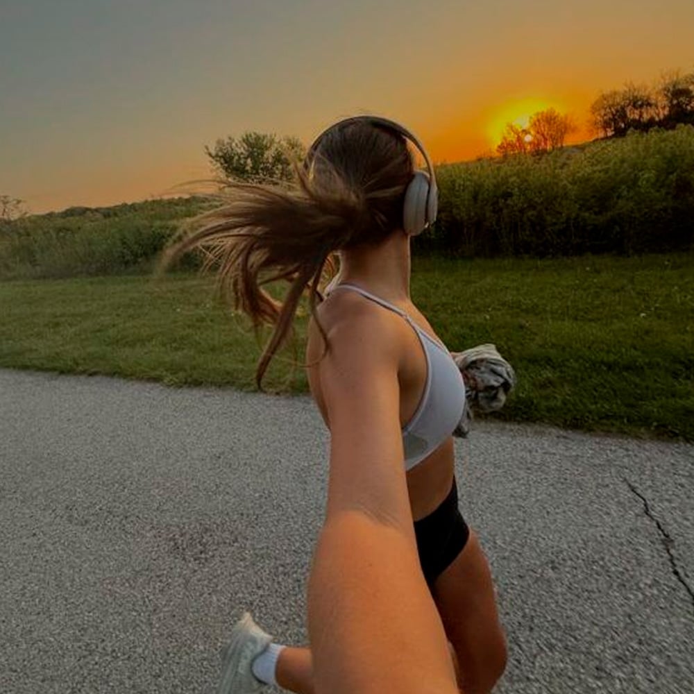 A woman in athletic wear runs on a path at sunset, wearing headphones and holding someone's hand behind her. Grassy fields and trees line the background under an orange sky.