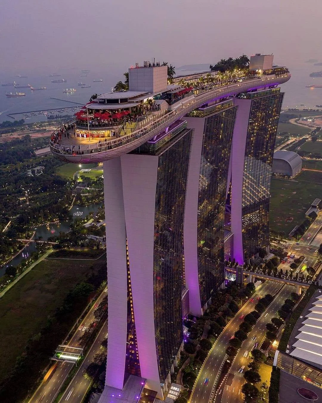 Aerial view of Marina Bay Sands in Singapore at dusk, with its three tall towers connected by a large rooftop sky park, glowing with purple lights and surrounded by cityscape and water.