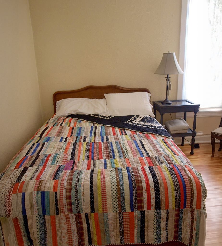 A neatly made bed with a colorful patchwork quilt, white pillows, and a black-and-white throw in a cozy bedroom. A bedside table with a lamp and books sits by a window letting in natural light.