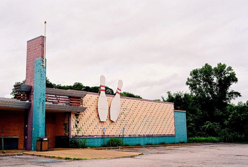 An abandoned bowling alley with faded pastel walls, boarded windows, and two large bowling pins as signage. The building is surrounded by overgrown vegetation and an empty, cracked parking lot under a cloudy sky.