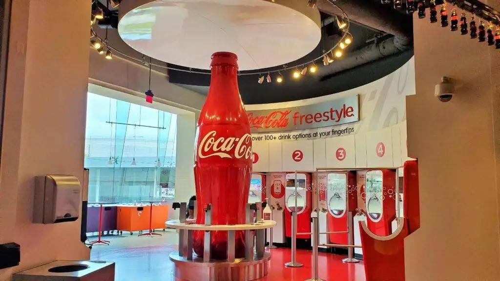 A large Coca-Cola bottle statue stands in the center of a modern soda fountain area with Coca-Cola Freestyle machines and a red and white color scheme. Bright lighting and windows create an inviting atmosphere.