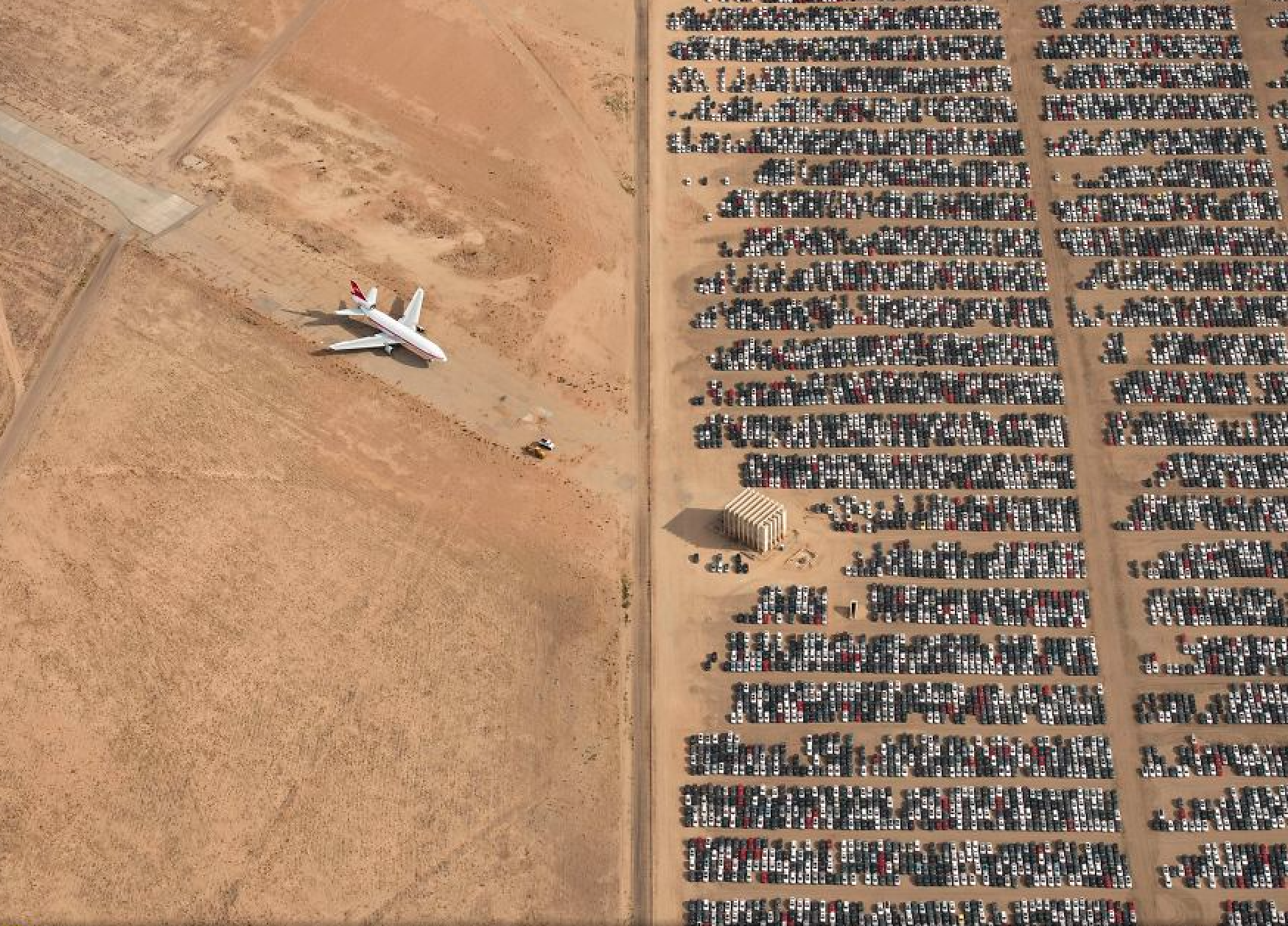 Aerial view of a large, dry, sandy area with a parked airplane on the left and rows of tightly packed cars on the right, separated by a clear dividing line.