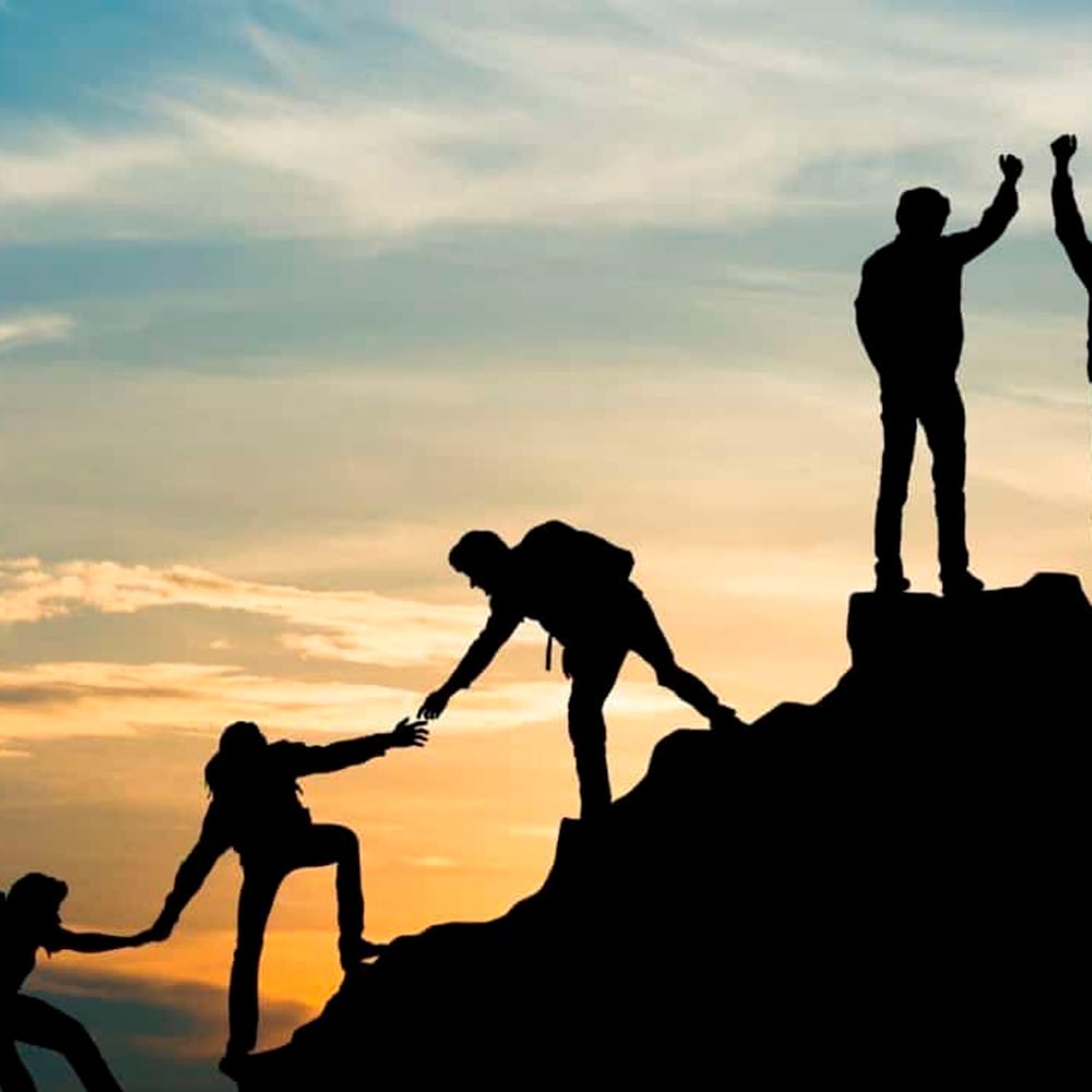 Silhouettes of people climbing a steep hill at sunset, with one person reaching down to help another up, symbolizing teamwork and support.