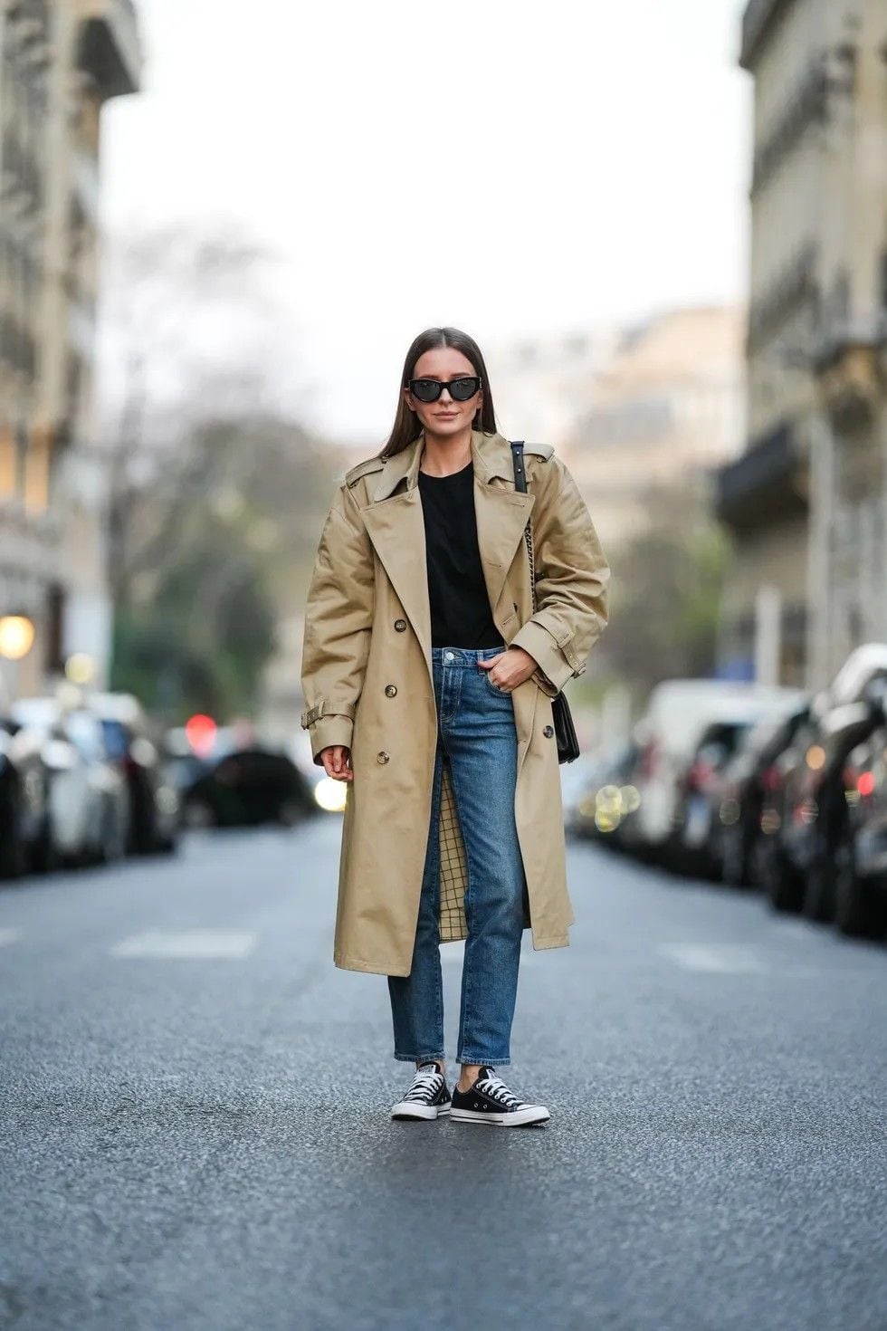 A person standing confidently on a city street wearing a tan trench coat, black shirt, blue jeans, black-and-white sneakers, and sunglasses, with parked cars and buildings on either side.