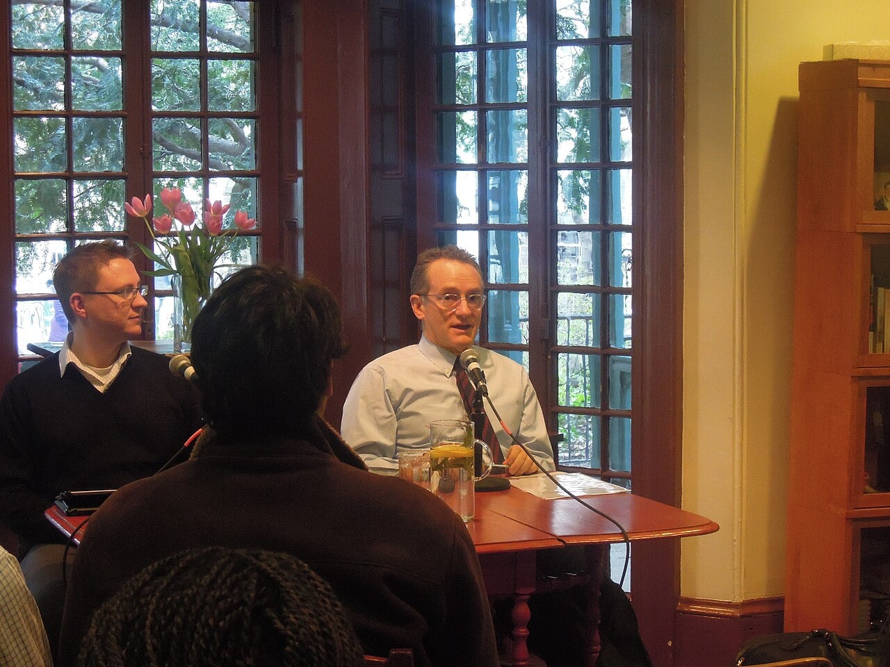 Two men sit at a wooden table with microphones, speaking to an audience in a room with large windows. One man is talking while the other listens. There is a vase of pink tulips and bookshelves in the background.
