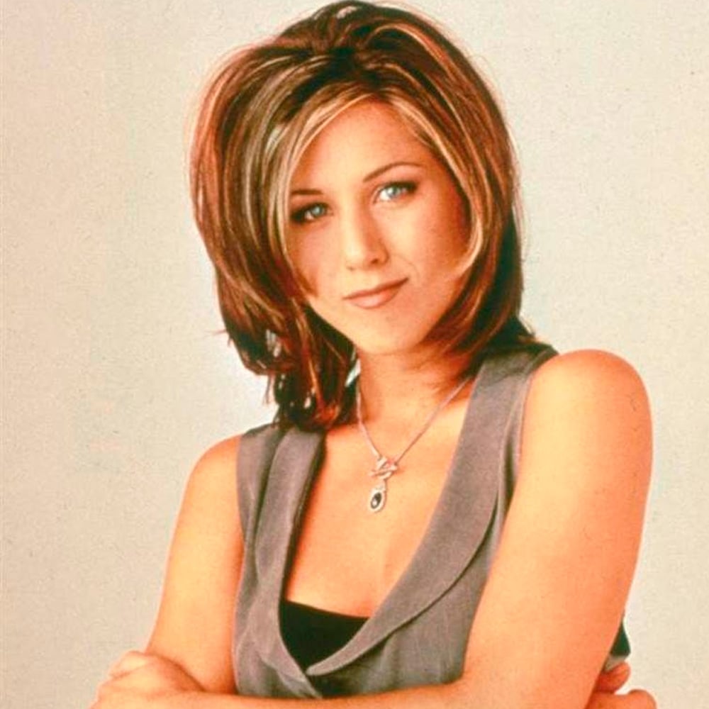 A woman with layered, shoulder-length brown hair and blonde highlights stands with her arms crossed, wearing a sleeveless gray vest over a black top and a pendant necklace, looking at the camera with a slight smile.