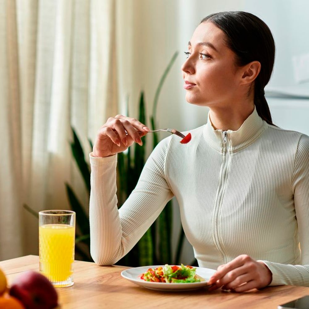 A woman in a white zip-up top sits at a table with a fork and a plate of salad. A glass of orange juice is beside her. She looks to the side, with bright light coming through sheer curtains behind her.