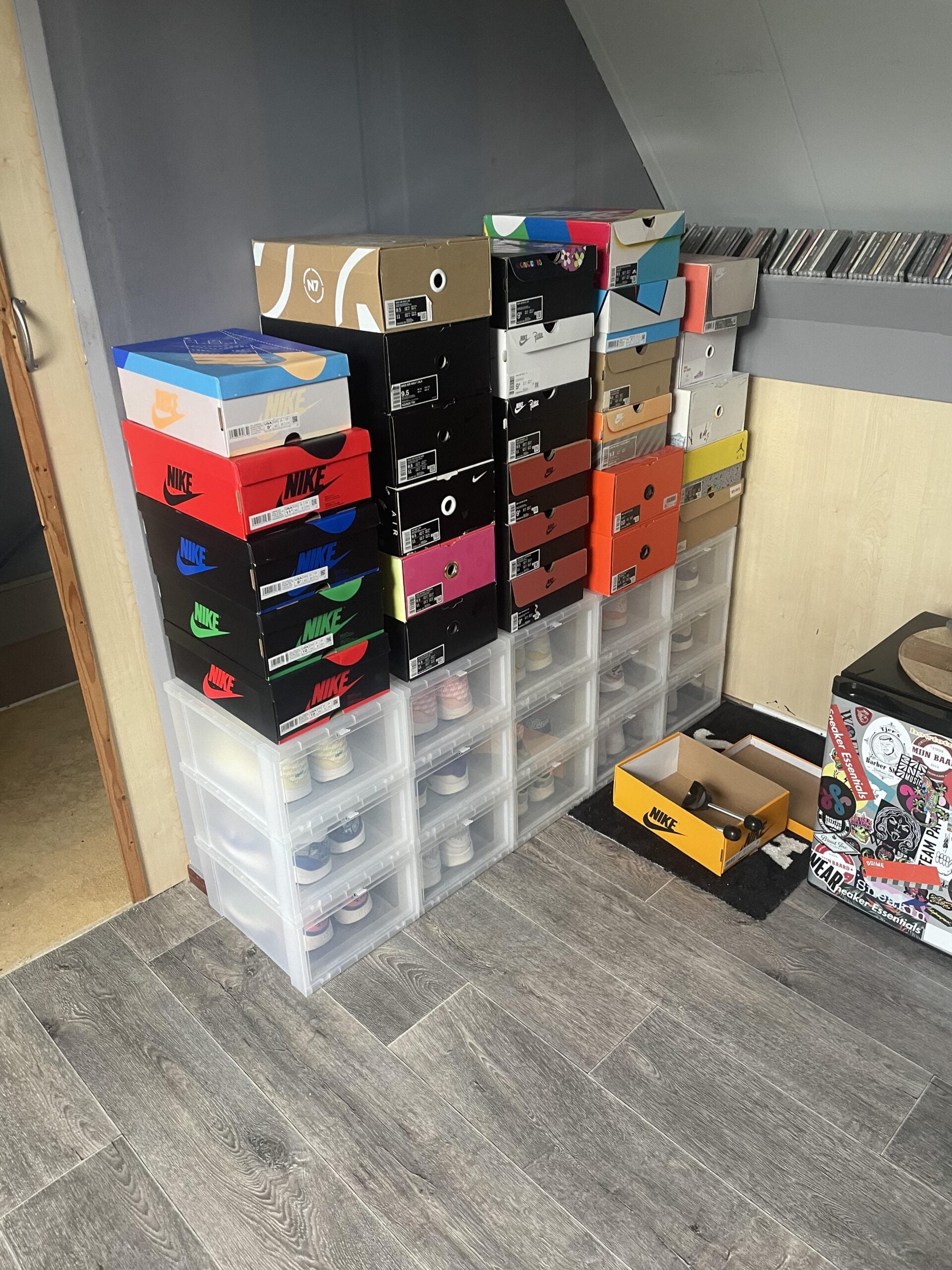 A stack of colorful sneaker boxes, mostly Nike and other brands, sit on top of clear plastic storage drawers in a room with gray wood flooring, next to a record player and shelves of CDs. An open box is on the floor.