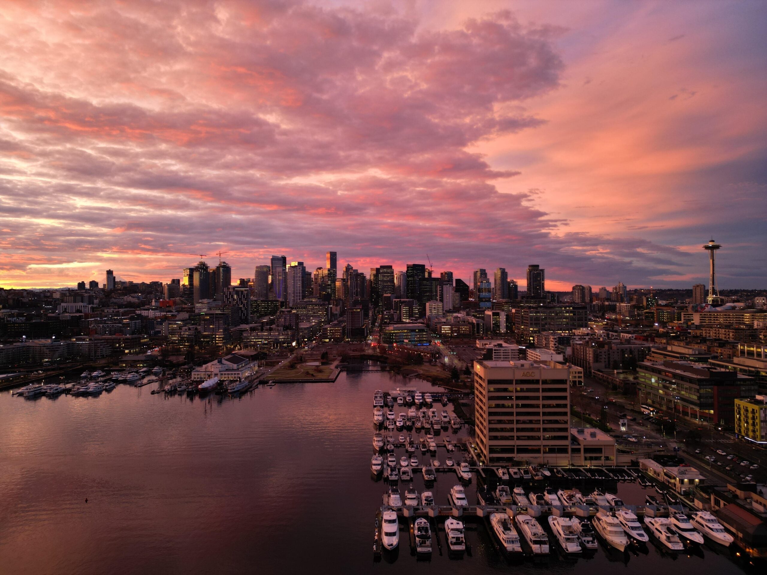 A city skyline at sunset with dramatic pink and orange clouds. High-rise buildings and the Space Needle are visible in the background. Marina with boats and calm water in the foreground.