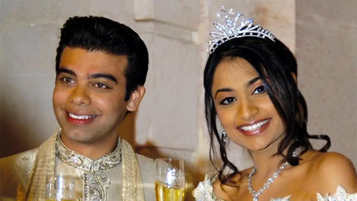 A smiling man in traditional attire and a woman wearing a tiara and white dress hold champagne glasses together, posing for a celebratory photo.