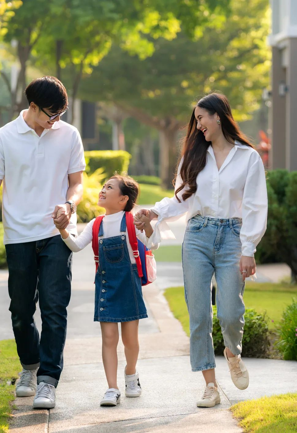 A smiling family of three, two adults and a young girl with a red backpack, walk hand in hand along a sunny sidewalk in a green, tree-lined neighborhood.