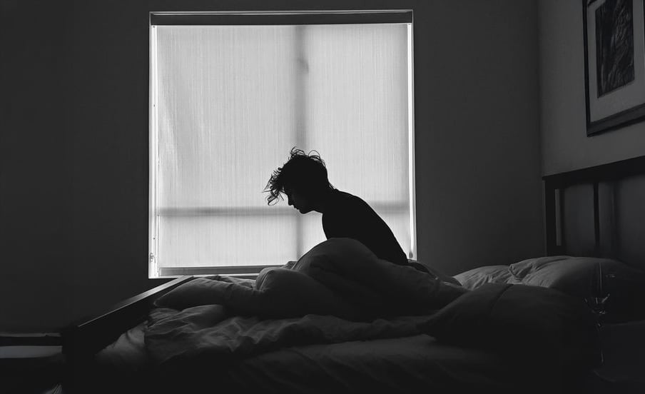 Silhouette of a person sitting on a bed in a dimly lit room, facing a large window with drawn blinds. The person appears thoughtful or tired, with messy hair and bedding around them.