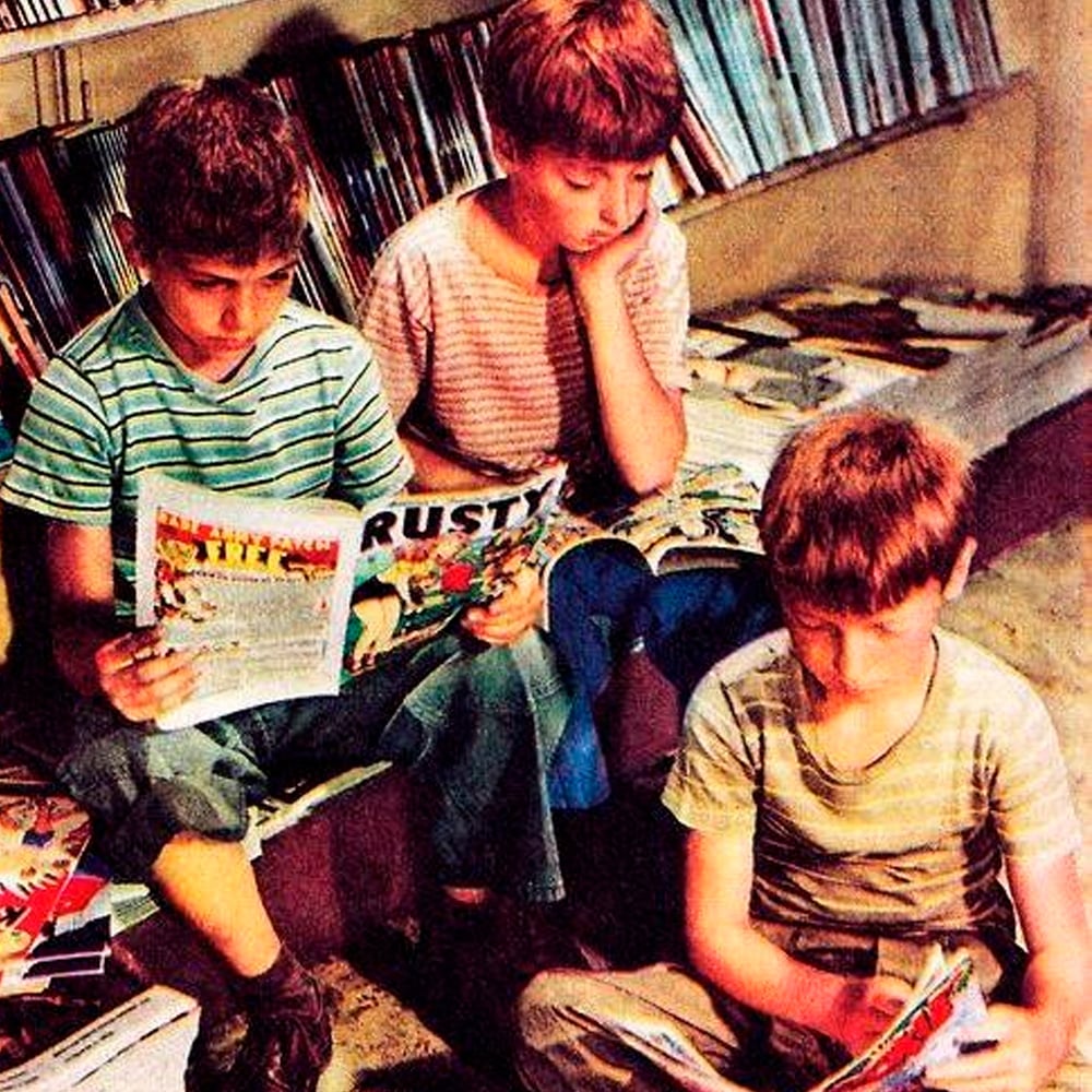 Three children sit on the floor and bench in a library or bookstore, reading comic books. Shelves filled with books surround them. All three wear striped shirts and appear focused on their reading.