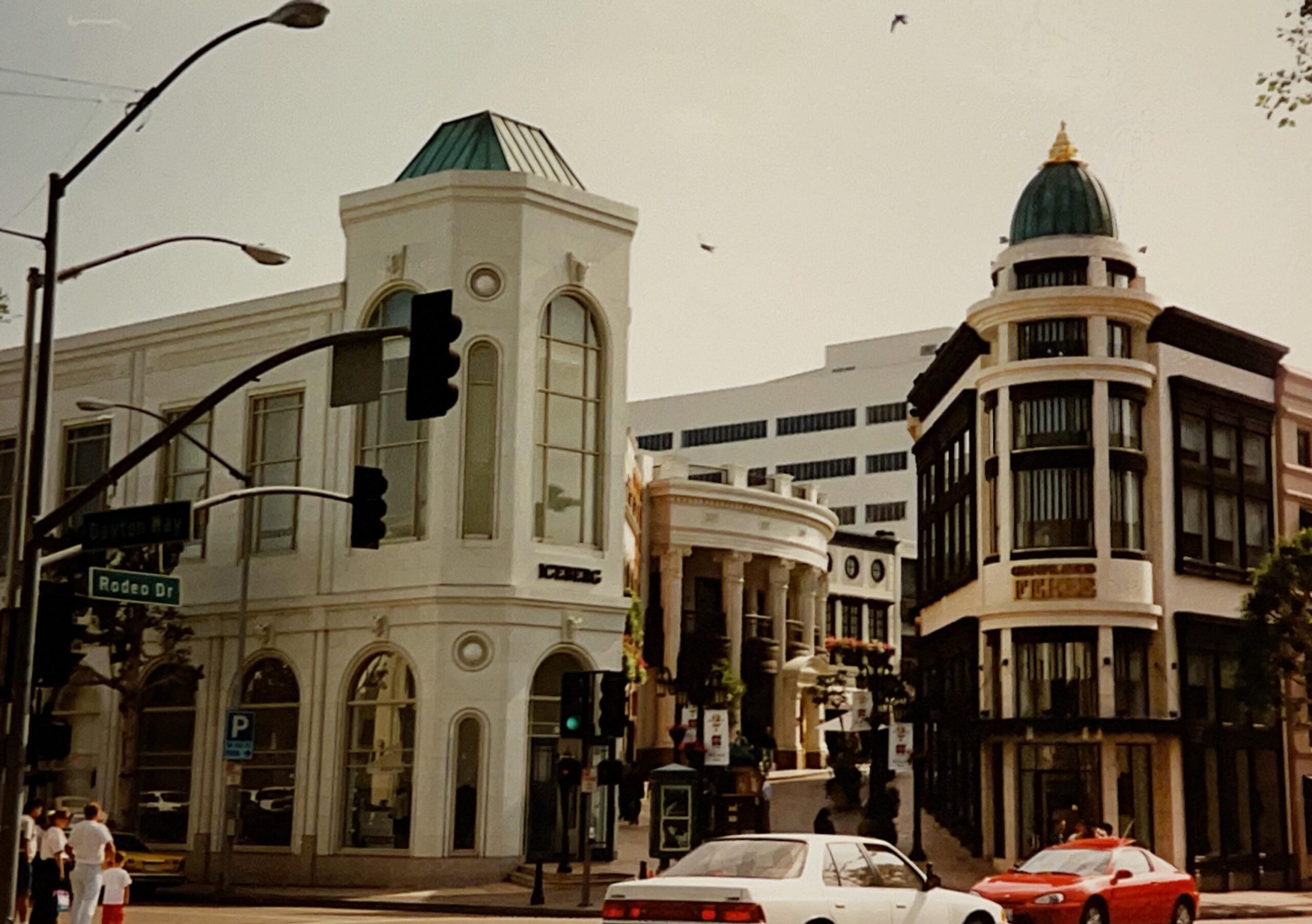 A sunny street corner with two ornate buildings at an intersection. One building is white with arched windows, the other black and white with a green dome. There are cars and pedestrians, and a street sign reads "Rodeo Dr.