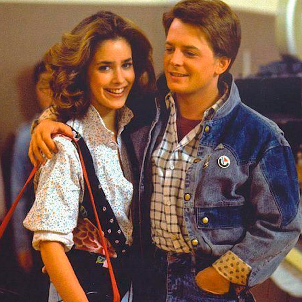 A young woman and man stand close together, smiling. The woman has long wavy hair and wears a patterned shirt with suspenders, while the man, in a denim jacket and plaid shirt, has his arm around her shoulders.