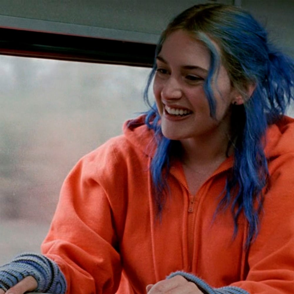 A woman with blue hair and an orange zip-up jacket smiles while sitting indoors, with a blurred window in the background.