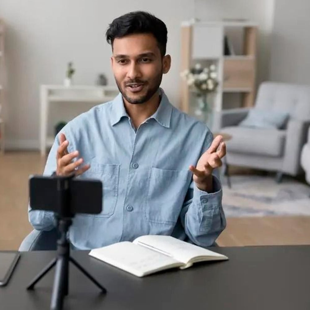 A man sits at a desk with an open notebook, speaking and gesturing towards a smartphone on a tripod, possibly recording or on a video call, in a bright, modern living room.