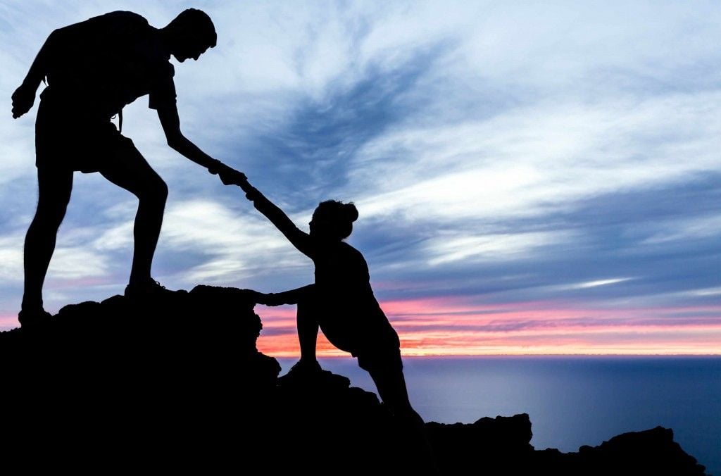 Silhouette of one person helping another climb a rocky hill at sunset, with a colorful sky and ocean in the background.