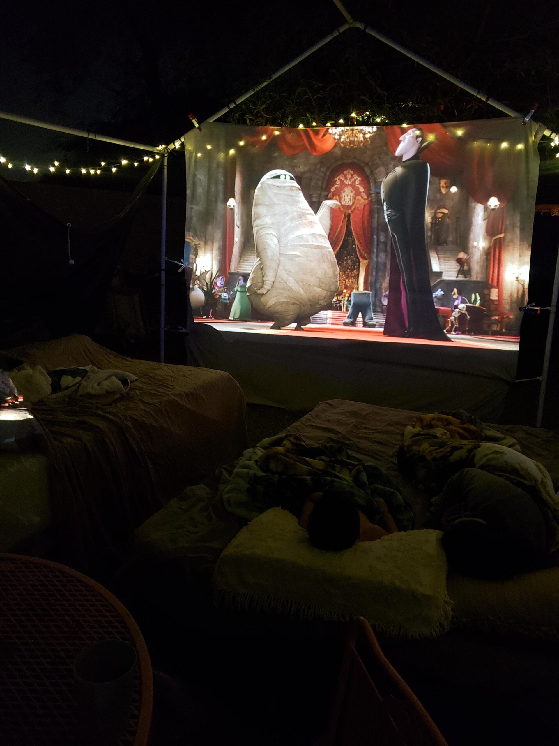An outdoor movie night setup with beds and blankets under string lights, showing an animated film on a projector screen. A child is lying down and watching the colorful movie scene.