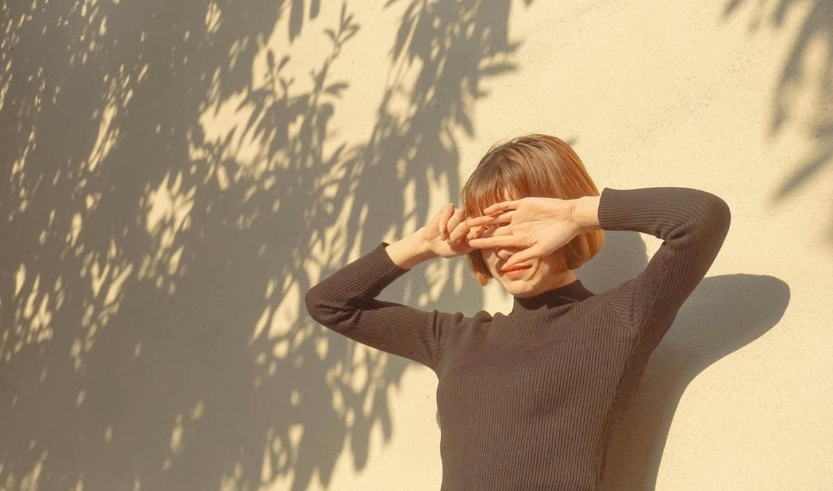 A person with short hair and a black long-sleeve shirt stands against a sunlit wall, shielding their eyes from the bright sunlight with their hands. Leaf shadows are cast on the wall beside them.