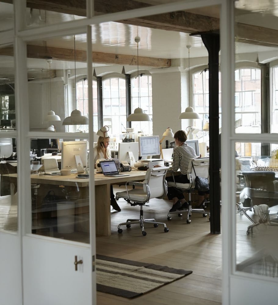 Modern open-plan office with large windows, wooden floors, and exposed beams. Several people are working at desks with computers. White chairs, pendant lights, and office supplies are visible in the bright workspace.
