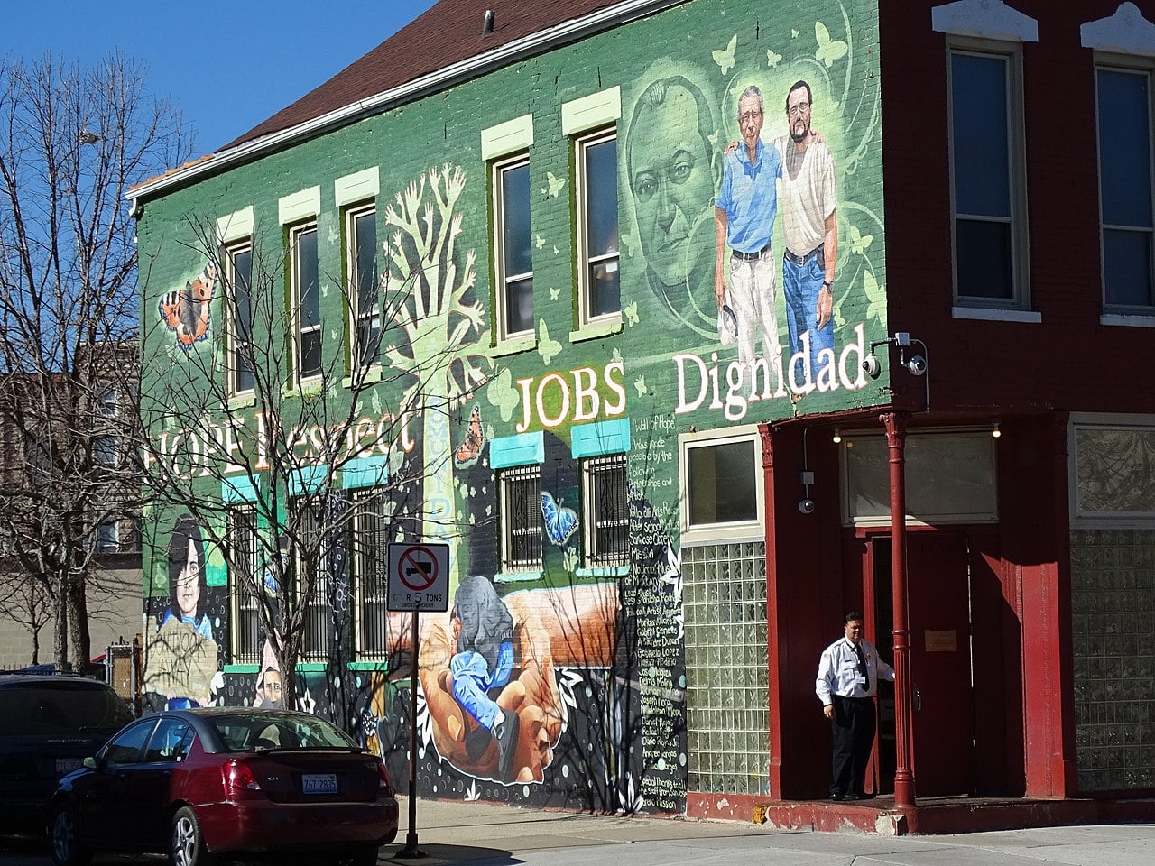A colorful mural on a building features images of people, a large tree, butterflies, and words like "Respect," "JOBS," and "Dignidad" in big letters. A man in a white shirt stands near the entrance. Cars are parked nearby.