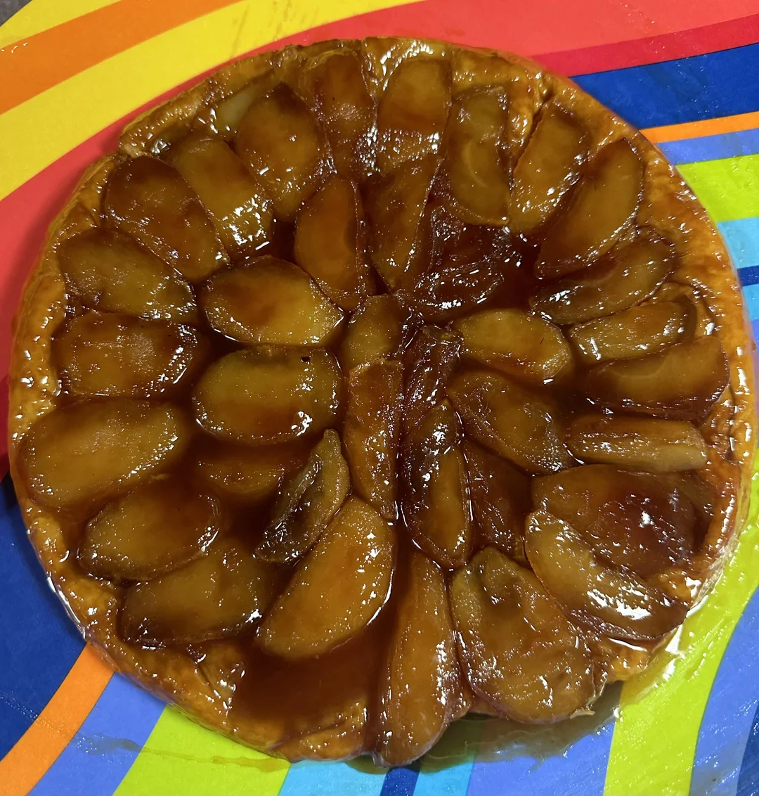 A caramelized upside-down apple tart, or tarte Tatin, with glossy sliced apples arranged in a spiral pattern, sits on a colorful striped surface with red, yellow, orange, blue, and green lines.