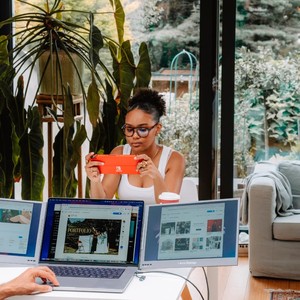 A woman sits at a table with three open laptops, holding a red Nintendo Switch. She wears glasses and is focused on the game. Large plants and a window with a view of a garden are in the background.