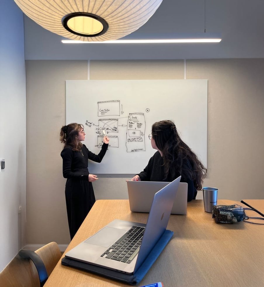 Two women are working in a modern office; one stands and points at diagrams on a whiteboard while the other sits at a table with laptops, listening attentively.